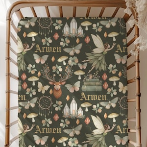 Puede incluir: Una sábana bajera ajustable verde oscuro con un patrón caprichoso. El diseño incluye hadas, mariposas, setas, ciervos y el nombre "Arwen" en una fuente decorativa. La sábana está colocada sobre una estructura de cuna de madera.