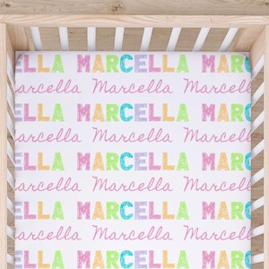 May include: A white crib sheet with the name "ELLA MARCELLA" in repeating rows. The name is in colorful, block letters: green, yellow, pink, and blue. The crib sheet is inside a light wood crib.