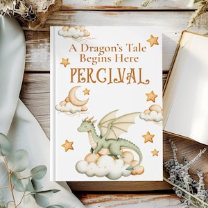 May include: A hardcover book titled "A Dragon's Tale Begins Here PERCIVAL" with a whimsical illustration of a green dragon on a cloud, surrounded by stars and a crescent moon. The book is set against a wooden background.