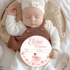 May include: A pink and white round wooden baby announcement plaque with a ballerina illustration and the text "Eloise Penrose" with spaces for the date, time, length, and weight.