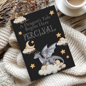 May include: A black book cover with the title "A Dragon's Tale Begins Here PERCIVAL". The cover features a gray dragon on clouds, with stars and a crescent moon. The book is on a white knitted blanket, with a cup of coffee in the background.