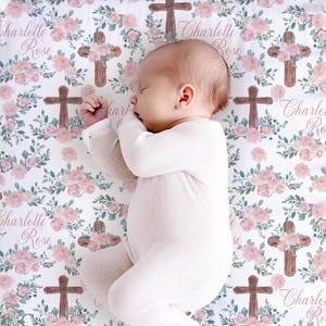 May include: A baby in a white onesie lies on a crib sheet with a floral and cross pattern. The sheet features pink roses, green leaves, and brown crosses. The name "Charlotte Rose" is written in cursive script on the sheet.