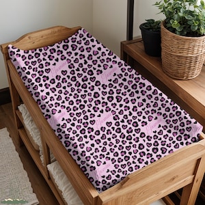 May include: A pink changing pad cover with a leopard print design. The pattern includes black and purple spots and heart shapes. The name "Jasmine" is printed in a cursive font across the pad.