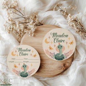 May include: Two circular wooden baby announcement plaques. One features the name "Meadow Claire" in cursive, with a snake illustration, moon, stars, and floral accents. The other has the same name and illustration, with spaces for date, time, length, and weight.