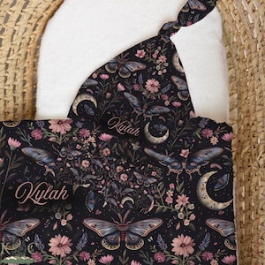 May include: A black baby hat and blanket set featuring a repeating pattern of moths, crescent moons, and pink flowers. The name Kylah is printed in a cursive script on both the hat and the blanket. The fabric has a dark background with botanical elements.