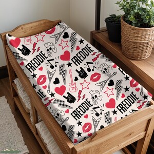 May include: A white changing pad cover with a repeating pattern of red hearts, stars, lightning bolts, guitars, skulls, and the name "FREDDIE" in black. The cover is on a wooden changing table.