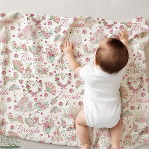 May include: A baby lies on a cream-colored blanket with a repeating pattern of pink and green illustrations. The design includes swans, bunnies, dresses, umbrellas, and the name "Cressida" in a script font. The blanket is rectangular.