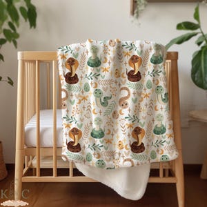 May include: A white baby blanket with a pattern of green and brown snakes, leaves, and flowers. The blanket is soft and cuddly, perfect for a nursery or crib.