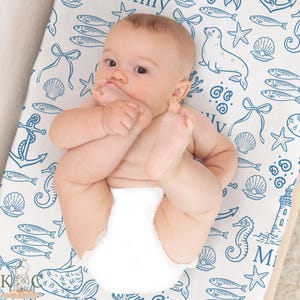 May include: A baby lying on a white changing pad with a blue nautical pattern of fish, seashells, anchors, and seahorses. The baby is wearing a white diaper.