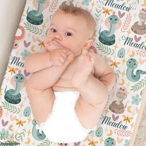 May include: A baby lies on a changing pad with a whimsical snake and floral pattern. The pad features a white background with teal, pink, and brown illustrations of snakes, dragonflies, and the word "Meadow". The baby wears a white diaper.