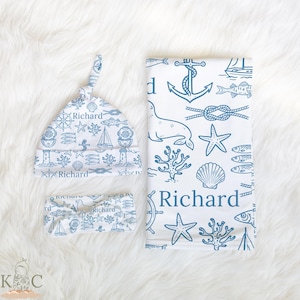 May include: A baby set with a nautical theme, comprising a knotted hat, headband, and blanket in white and blue. The set is decorated with illustrations of anchors, sailboats, fish, and seashells. The name "Richard" is printed on the items.