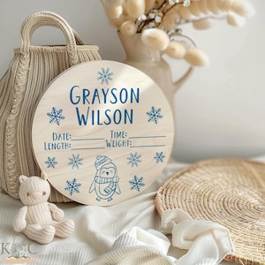 Puede incluir: Un letrero redondo de madera con el nombre "Grayson Wilson" en azul, rodeado de copos de nieve y una ilustración de pingüino. Incluye espacios para fecha, hora, longitud y peso. Un oso de peluche tejido y una bolsa de mimbre en el fondo.