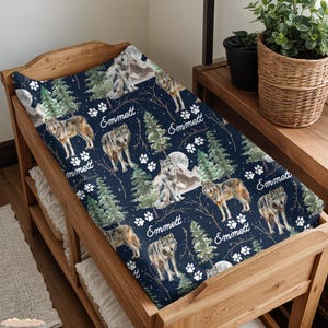 Peut inclure: Une housse de matelas à langer bleu marine avec un motif répété de loups, de pins, d'empreintes de pattes et de montagnes. Le nom "Emmett" est écrit en blanc. La housse est sur une table à langer en bois.
