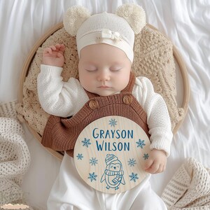Puede incluir: Un bebé dormido con un gorro blanco de punto con pompones y un mono marrón. El bebé está acostado en una cesta de madera sobre una superficie blanca. Un letrero redondo de madera con el nombre "Grayson Wilson" y una ilustración de pingüino.