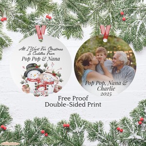 May include: Two round Christmas ornaments with red ribbon hangers. One features a snowman family with the text "All I Want For Christmas Is Cuddles From Pop Pop & Nana." The other shows a family photo with the text "Pop Pop, Nana & Charlie 2025."
