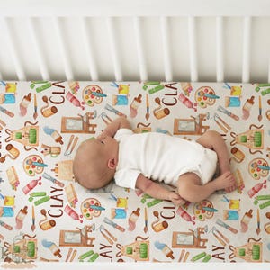 May include: A baby lies on a cot sheet patterned with art supplies. The design features paint palettes, paint tubes, brushes, and the word "CLAY" in brown. The sheet is white with a watercolour style design.