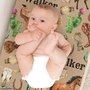 Peut inclure: Un bébé en couche est allongé sur un matelas à langer beige sur le thème du cow-boy. Le matelas présente des illustrations à l'aquarelle de cactus, de chevaux, de bottes et d'une guitare. Le nom "Walker" est imprimé.