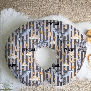 May include: A gray and white striped nursing pillow with a dragon, moon, and star pattern. The name "PERCIVAL" is printed on the pillow. The pillow is designed for comfort and support during feeding.