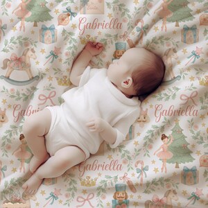 May include: A baby wearing a white onesie lies on a patterned blanket. The blanket features a Christmas-themed design with illustrations of ballerinas, Christmas trees, and rocking horses. The name "Gabriella" is printed on the blanket.
