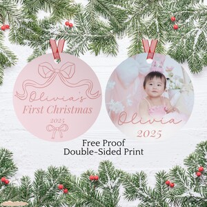 May include: Two pink Christmas baubles with red ribbon hangers. One bauble has a bow design and the text "Olivia's First Christmas 2025". The other bauble has a photo of a baby and the name "Olivia" and the year "2025".