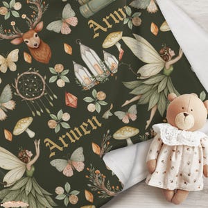 May include: A dark green baby blanket with a whimsical pattern featuring a deer, fairies, butterflies, mushrooms, and the name "Arwen" in gold lettering. A teddy bear wearing a floral dress is in the foreground.