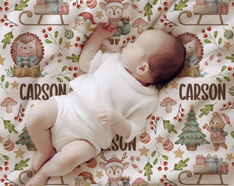 May include: A baby rests on a white blanket adorned with a Christmas pattern. The design features a deer, hedgehog, Christmas tree, gifts, and the name "CARSON". The blanket has a festive, whimsical design with red, green, and brown accents.