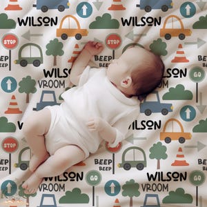 May include: A white baby blanket with a colorful print of cars, stop signs, arrows, and the name "Wilson" repeated throughout. The blanket is made of soft, comfortable fabric and is perfect for a baby boy.