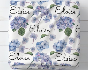 Op de afbeelding: Witte stof met een herhalend patroon van blauwe hortensia bloemen en de naam "Eloise" geschreven in zwarte cursieve letters.