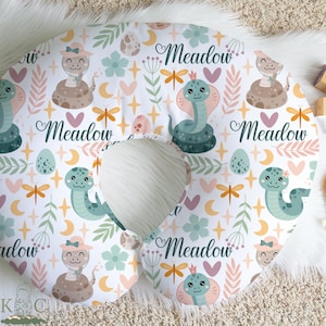 Pode incluir: Uma almofada de amamentação branca para bebé com um padrão fantasioso com cobras de desenho animado, ovos, flores e a palavra "Meadow". As cobras são em tons de verde, castanho e rosa, com o texto em itálico.