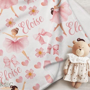May include: A white baby blanket with a pink and white ballerina pattern. The blanket features pink bows, cherry blossoms, and the name "Eloise" written in pink cursive. A brown teddy bear with a white lace collar and a floral dress is sitting on the blanket.