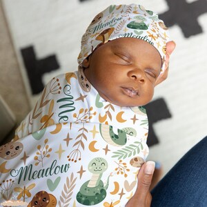 May include: A white swaddle blanket with a green snake, stars, and moons pattern. The blanket has the name "Meadow" written on it in green. A baby is wearing a matching hat with the name "Meadow" written on it.