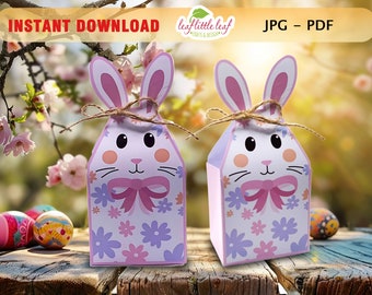 Puede incluir: Dos cajas de regalo de conejo de Pascua imprimibles con orejas rosas, un lazo rosa y un patrón floral. Las cajas están sobre una superficie de madera con huevos de Pascua en el fondo. JPG - PDF.