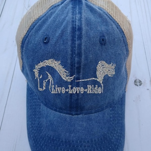 May include: A blue denim trucker hat with a beige mesh back. The hat features a silver glitter horse design and the text "Live-Love-Ride".