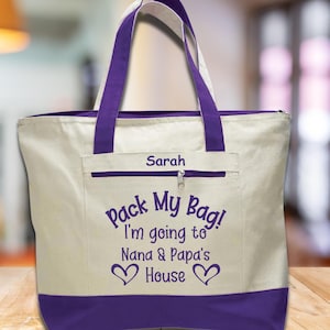 May include: A white canvas tote bag with purple handles and trim. The bag has a zippered pocket on the front and the text "Pack My Bag! I'm going to Nana & Papa's House" with hearts on either side. The name "Sarah" is printed above the text.