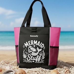 May include: Black tote bag with pink side panels and black handles. The bag features the text "Mermaid Stuff" in white, along with a mermaid graphic. The name "Kennedy" is printed above the graphic. The bag is on a sandy beach.