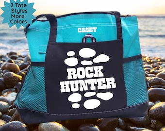 May include: A teal tote bag with a black front pocket featuring the words "ROCK HUNTER" in white. The bag has black handles and side mesh pockets. The name "CASEY" is printed in white on the teal top. Additional text reads "2 Tote Styles More Colors".