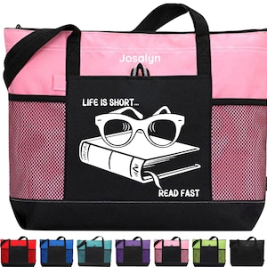 May include: A pink and black tote bag with a white design that says "Life is short... Read fast" and features a book with sunglasses on top. The bag has a mesh pocket on the side and a black zippered pocket on the front.