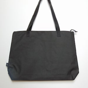 Op de afbeelding: Een zwarte canvas tote bag met twee zwarte handvatten en een ritssluiting.