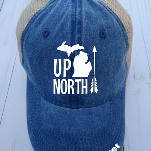 May include: A blue and tan trucker hat with a white outline of the state of Michigan and the words "UP NORTH" with an arrow pointing up. The name "Juliet" is embroidered on the bottom of the hat.