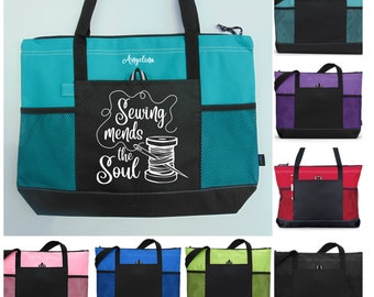 May include: A turquoise tote bag with a black bottom and a zippered top. The bag has a large pocket on the front with the text "Sewing mends the soul" and a drawing of a spool of thread and needle. The bag has a mesh pocket on the side. The bag is shown in multiple colors, including teal, purple, red, pink, blue, green, and black.