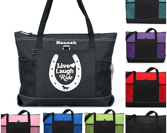 May include: A black tote bag with a white horseshoe design and the text "Live Laugh Ride". The bag has a zipper closure and a mesh pocket on the side. The bag is personalized with the name "Hannah". The bag is shown in multiple colors, including teal, purple, red, pink, blue, green, and black.