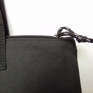 Op de afbeelding: Zwarte tote bag met een ritssluiting en een trekkoord aan de bovenkant. De tas heeft een zwarte band en een witte achtergrond.