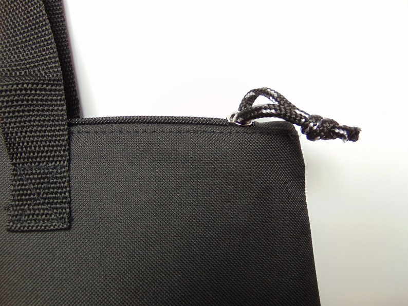 Op de afbeelding: Zwarte tote bag met een ritssluiting en een trekkoord aan de bovenkant. De tas heeft een zwarte band en een witte achtergrond.