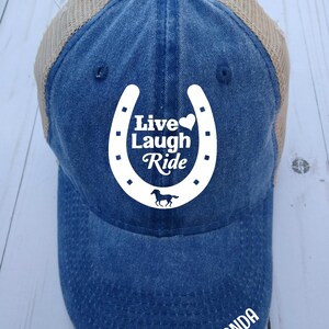 May include: A blue denim and beige mesh trucker hat with a white horseshoe design that says "Live Laugh Ride" and a small white horse silhouette. The hat has the name "Amanda" embroidered on the back.
