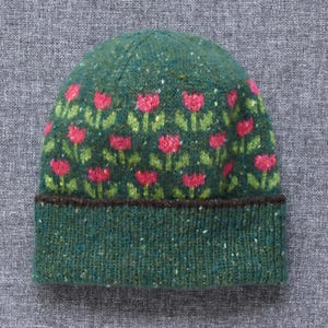May include: A dark green knit beanie featuring a repeating pattern of pink tulips with green stems and leaves. The hat has a wide, ribbed cuff at the base and is crafted from a speckled yarn that includes small flecks of lighter green and white.