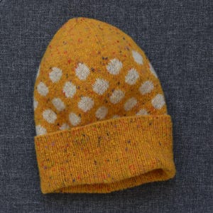 Puede incluir: Un gorro de punto color amarillo mostaza con un patrón repetitivo de lunares blancos. El tejido tiene una textura moteada con pequeñas motas de varios colores en el hilo. El gorro incluye un borde ancho doblado en la parte inferior.