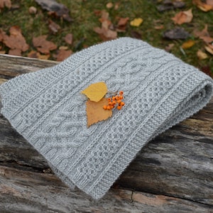 May include: A gray knitted scarf with a cable knit pattern. The scarf is folded and has a few yellow leaves and orange berries on top.