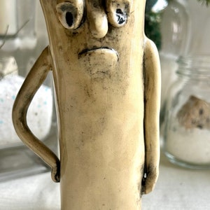 Könnte beinhalten: Eine Keramiktasse mit handbemaltem Gesicht. Die Tasse hat eine hellbraune Glasur und einen leicht abgenutzten Look. Das Gesicht hat einen traurigen Ausdruck mit großen Augen und einem heruntergezogenen Mund.