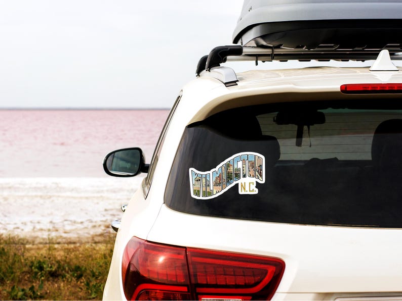 May include: White car with a sticker on the back window that says "WILMINGTON N.C." in a colorful font. The sticker is made up of images of buildings and other landmarks. The car is parked in front of a body of water with a pink hue.