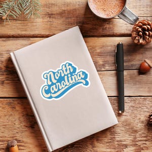 May include: A white sticker with blue text that says "North Carolina" in a retro font style. The sticker is on a white notebook.